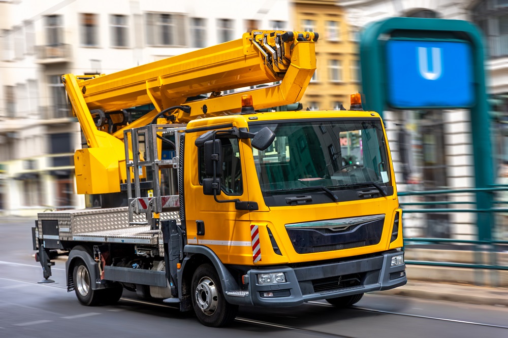 A bright yellow utility truck with a hydraulic lift drives through the city, representing maintenance, technology, and urban development.